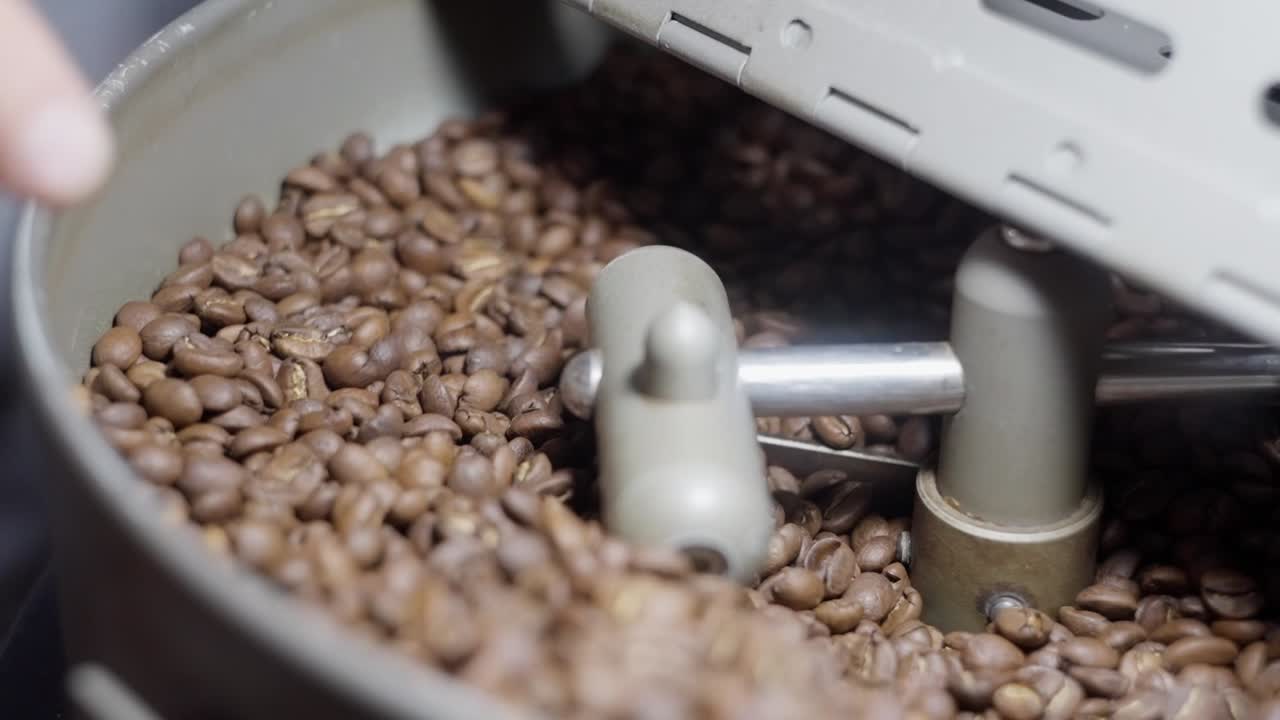 Close up view in slow motion of a coffee toaster with brown beans being toasted and a hand checking the process.