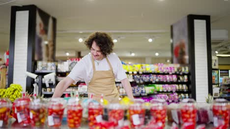 a confident guy with curly brunette hair in a yellow apron takes inventory of products and rearranges citrus fruits during his work in a supermarket