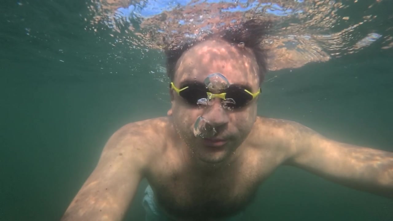 Uomo con occhialini da nuoto gialli è sott'acqua in acqua verde limpida. Sta espirando bolle, guardando verso la fotocamera.