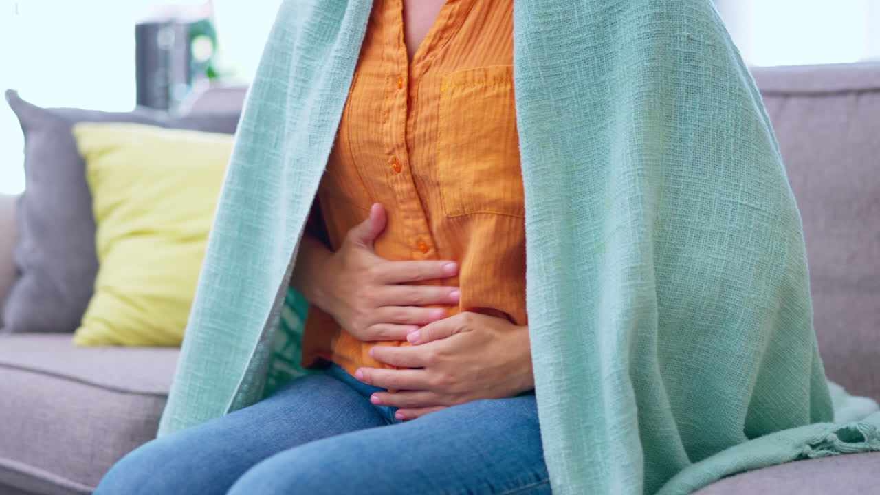 Hands, stomach pain and woman on sofa in house