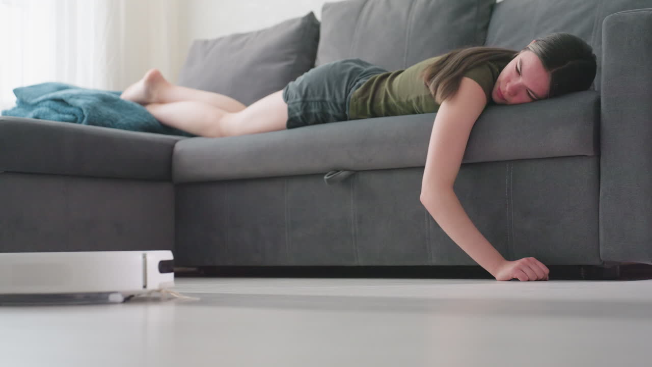 Young lady lies on gray couch lifting hand as robot vacuum approaches across bright living room floor, illustrating smart cleaning technology, relaxed lifestyle, and modern home