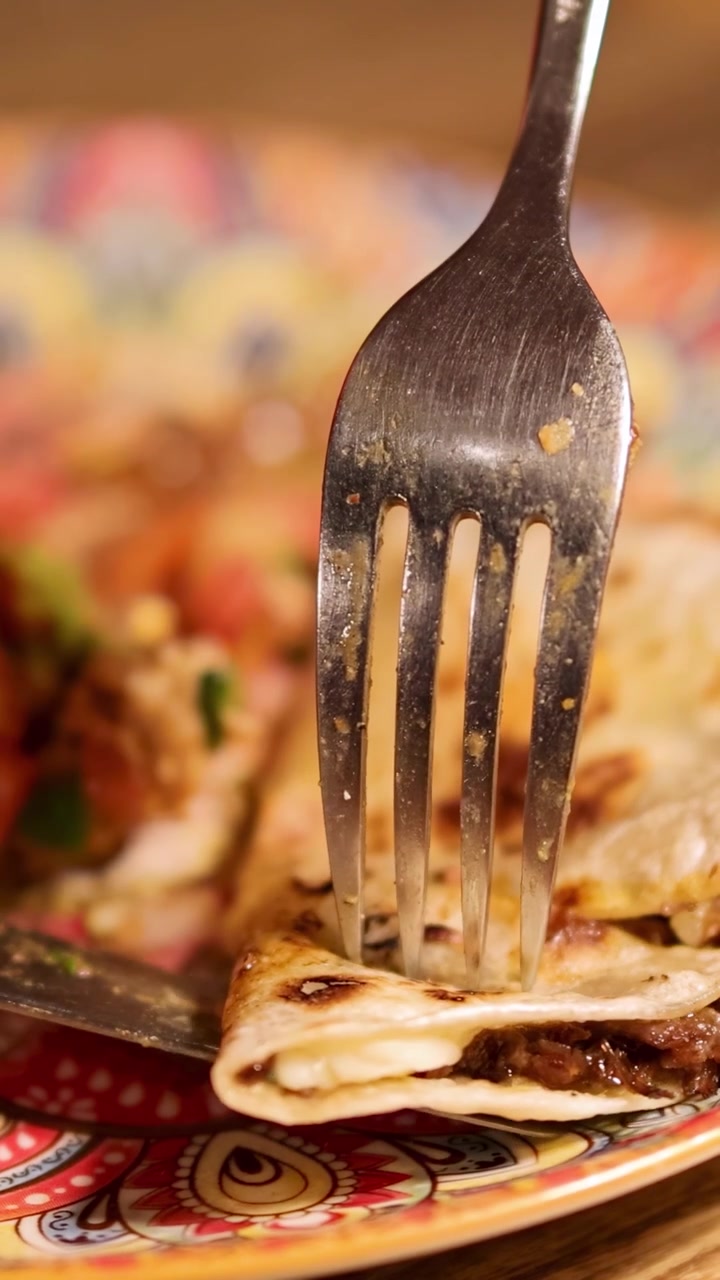 Detail vidličky a nože krájících hovězí fajitas na barevném talíři. Teplé osvětlení zvýrazňuje lákavou scénu.