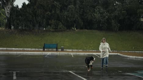 Glückliche-Blonde-Frau,-Die-Mit-Ihrem-Großen-Weißen-Hund-Davonläuft,-Während-Sie-Bei-Starkem-Regen-Im-Park-Spazieren-Geht