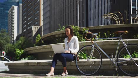 mujer afroamericana con bicicleta usando computadora portátil bebiendo café en el parque de la ciudad