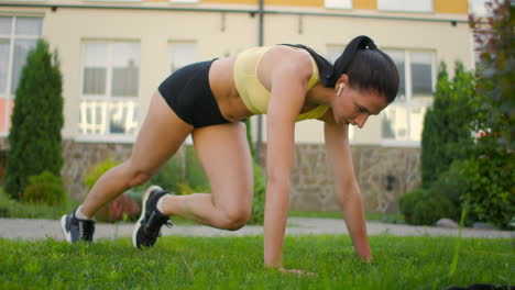 a woman with headphones trains standing on the grass in a plank performing a rock climber exercise in a slow-motion scheme in a city park