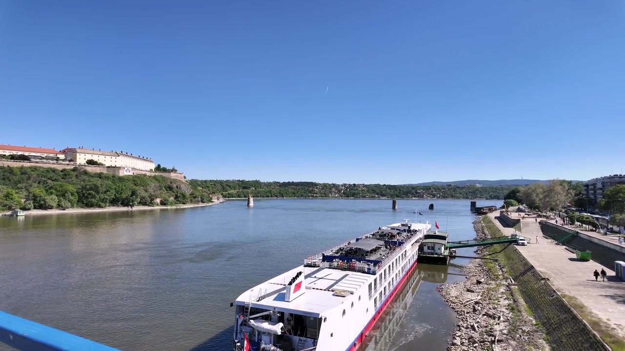 O barco de cruzeiro fluvial está saindo do porto em um dia ensolarado. Pessoas estão presentes perto da água e colinas cercam a área. O céu está limpo enquanto o cruzeiro inicia sua jornada ao longo do rio.