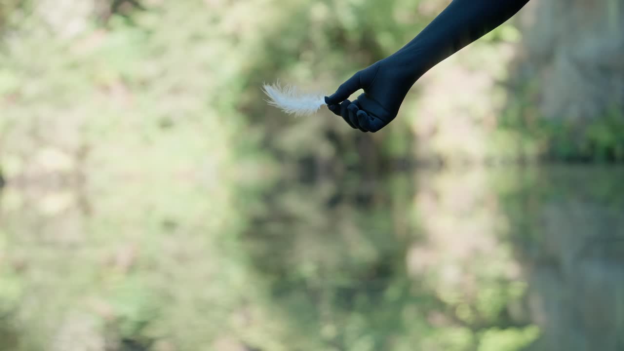 A woman's hand in a black glove drops a white feather onto the surface of the water. Natural background