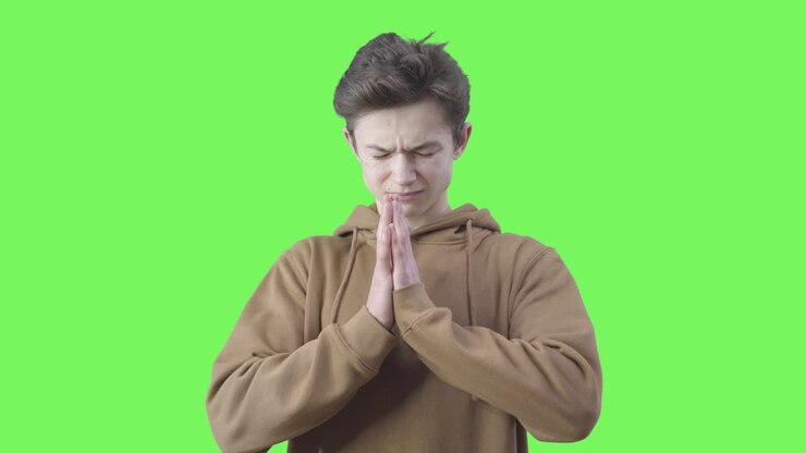 Ragazzo disperato che prega su sfondo verde. Ritratto di adolescente credente caucasico bruno con le mani giunte in preghiera. Fiducia, religione, fede. Chromakey