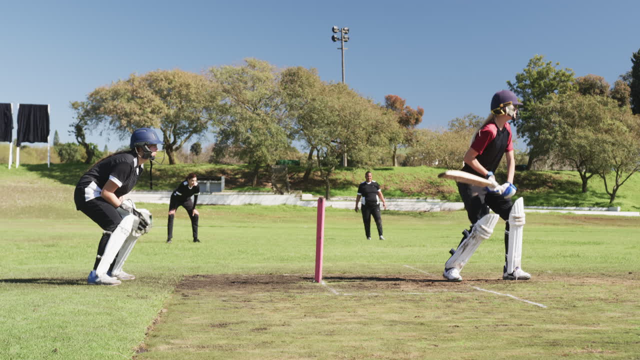 Playing cricket, female batter hitting ball while wicketkeeper and fielders watch