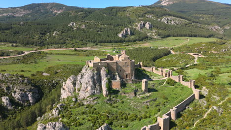 drone footage flying backward revealing an ancient medieval fortress on a rocky hilltop, surrounded by pine forest, farmlands, and distant mountain peaks under clear skies