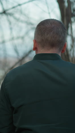 blurred close-up of a man in a green jacket outdoors with a background of tangled branches and distant scenery, with someone walked by and touch the tree