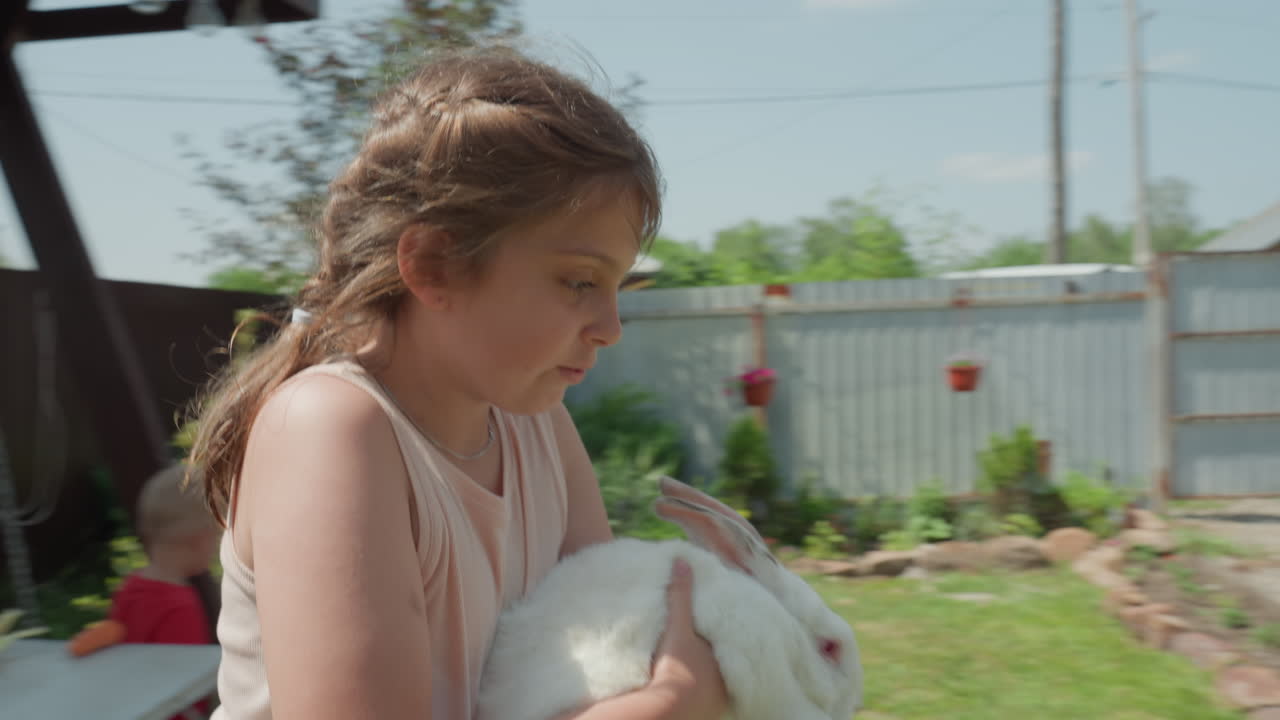 Young Girl Cradling White Rabbit Outdoors, Gentle Backyard Scene With Playground, Braided Hair, Sunlit Grass And Wooden Fence Child Holds Pet Protectively, Soft Movements And Warm Summer Mood