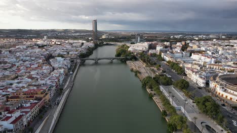 Aerial-dolly-along-iconic-Spain-Seville-river-and-bridge,-storm-clouds-behind