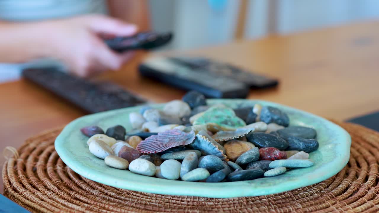 A hand reaches for and picks up a remote control from a wooden table, beside a decorative plate of assorted stones, in soft natural lighting