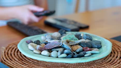 a hand reaches for and picks up a remote control from a wooden table, beside a decorative plate of assorted stones, in soft natural lighting