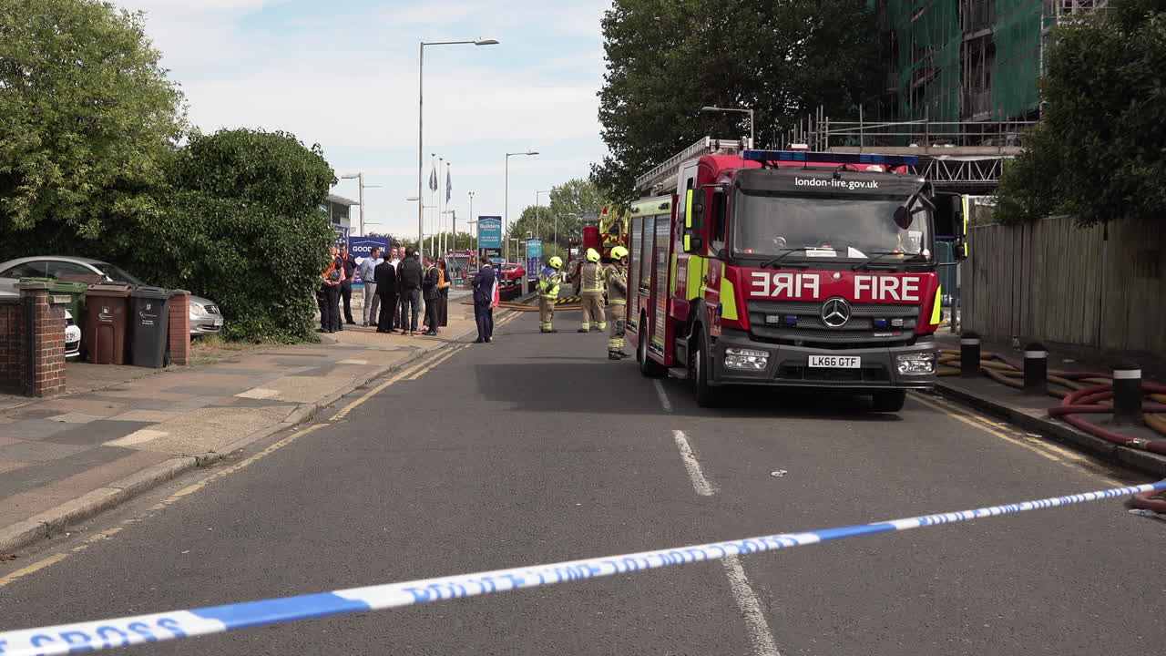 Police cordon tape flutters in the wind in front of firefighters and fire engines parked next to a tower block covered in flammable cladding that was destroyed by a fire.