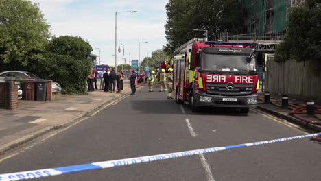 police cordon tape flutters in the wind in front of firefighters and fire engines parked next to a tower block covered in flammable cladding that was destroyed by a fire.