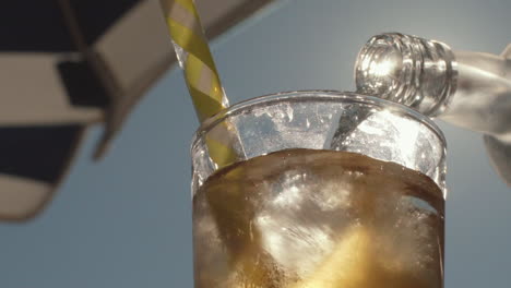 Close-up-of-transparent-liquid-being-poured-from-bottle-into-glass,-Backlit-with-sun-under-umbrella,-Slowmo