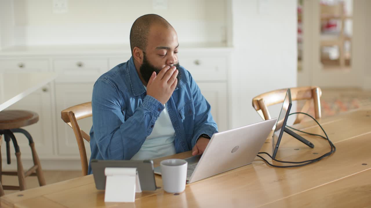 Man wearing denim typing at laptop at kitchen table, glancing at monitor, rubbing eyes from fatigue