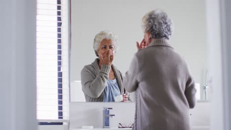 Mujer-Mayor-Afroamericana-Aplicando-Crema-Facial-Mientras-Se-Mira-En-El-Espejo-En-Casa