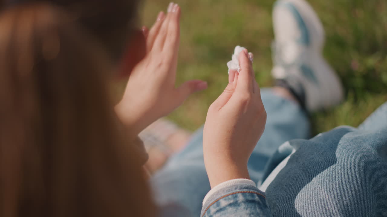 Female in denim cleans her hands thoroughly. Girl in outdoor sunlight carefully disinfects her hands. Young woman wearing denim jacket diligently refreshes her hands after enjoying snack outdoors