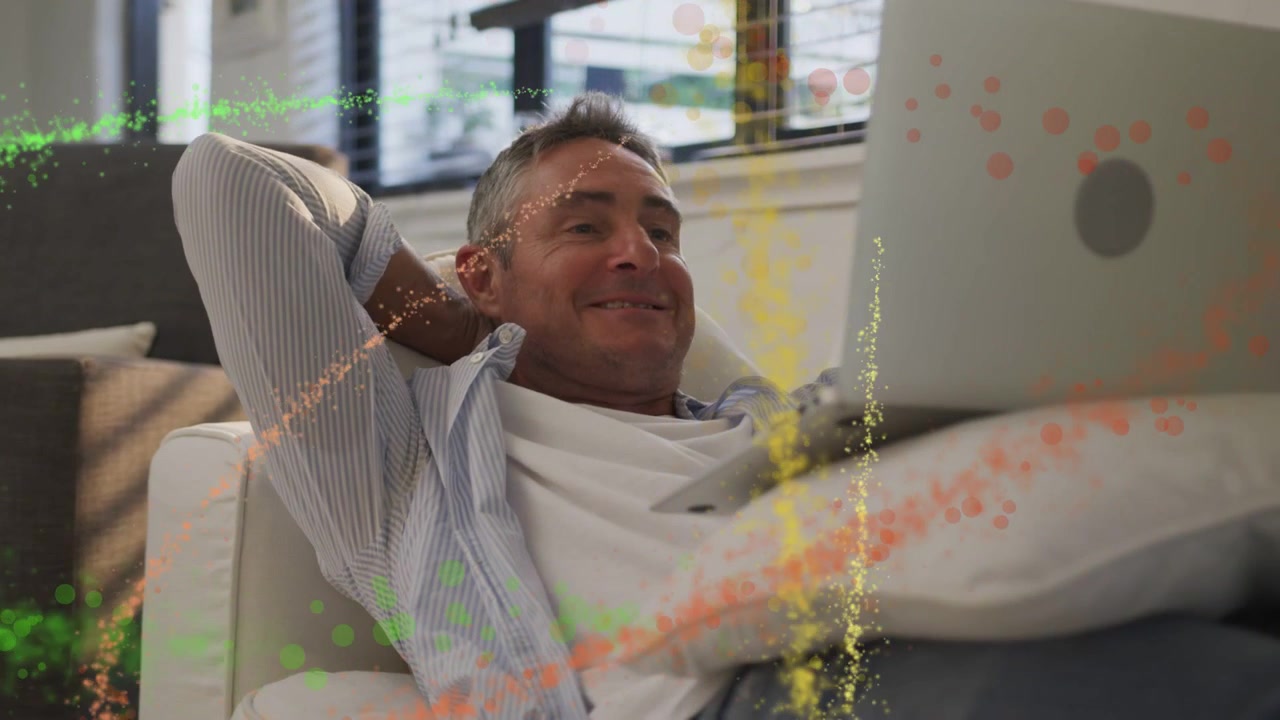 Mature man tapping laptop leaning back relaxing watching media as particles sweeping over him