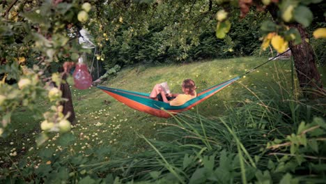 Man-walking-towards-and-laying-in-a-comfortable-hammock-tied-between-two-trees,-reading-a-book-surrounded-by-nature