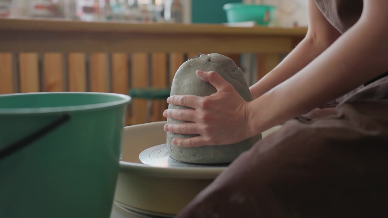 Giovane studente bianco modella l'argilla al tornio in un laboratorio di ceramica, le mani stabilizzano il blocco mentre è seduto su uno sgabello, con scaffali di smalti sullo sfondo, in un'atmosfera di apprendimento concentrato e con l'energia di una lezione pratica.