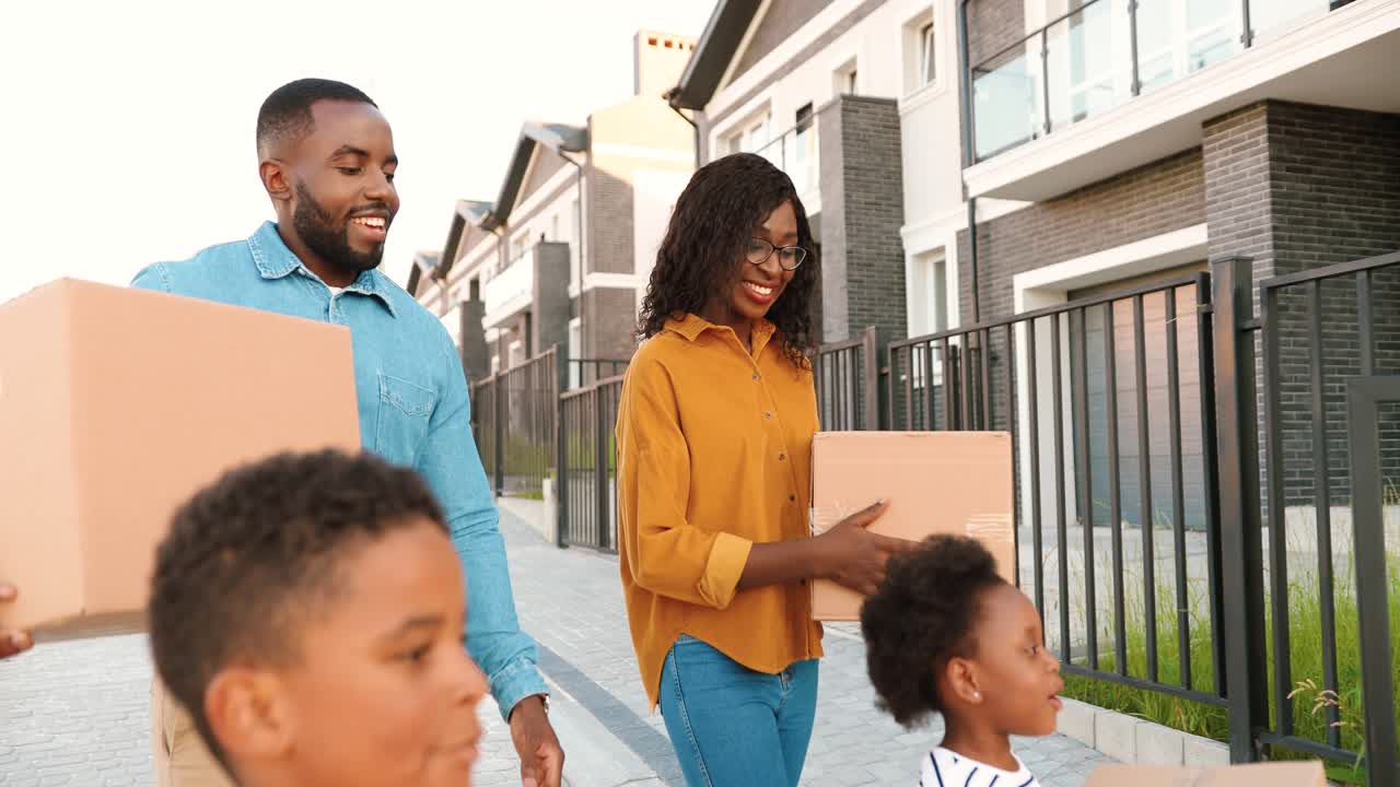Happy African American parents with children moving in new home and carrying carton boxes