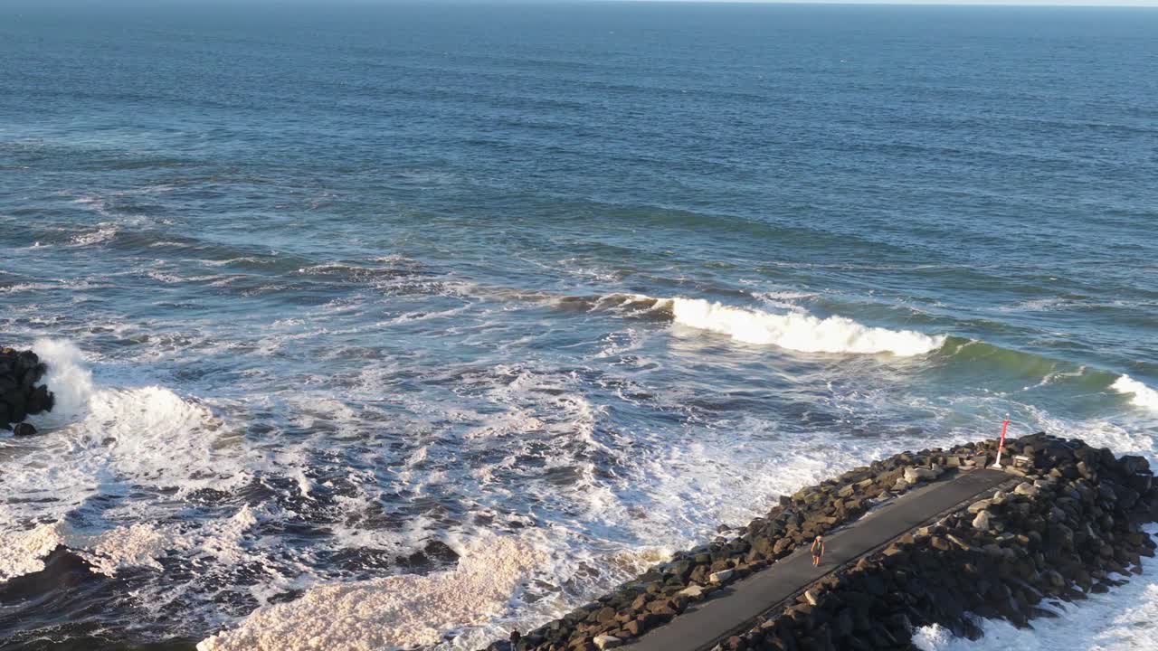 Aerial view of ocean waves hitting a rocky jetty under clear skies, showcasing dynamic water movement and coastal scenery