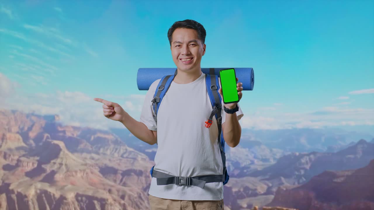 Asian Male Hiker With Mountaineering Backpack Showing Mock Up Green Screen Smartphone And Pointing To Side While Traveling At The Top Of Mountain