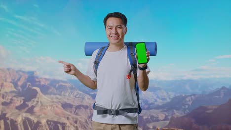 asian male hiker with mountaineering backpack showing mock up green screen smartphone and pointing to side while traveling at the top of mountain