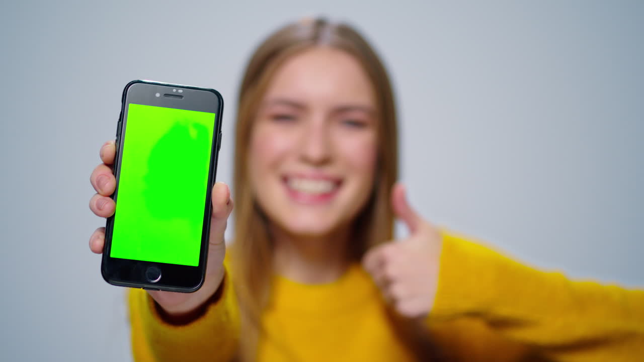 mujer sonriente mostrando un teléfono inteligente con pantalla verde en el estudio. niña pulgares hacia arriba