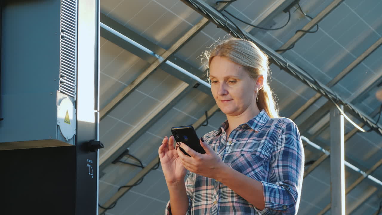 The Woman Uses A Smartphone Stands At The Inverter Under The Panels Of The House