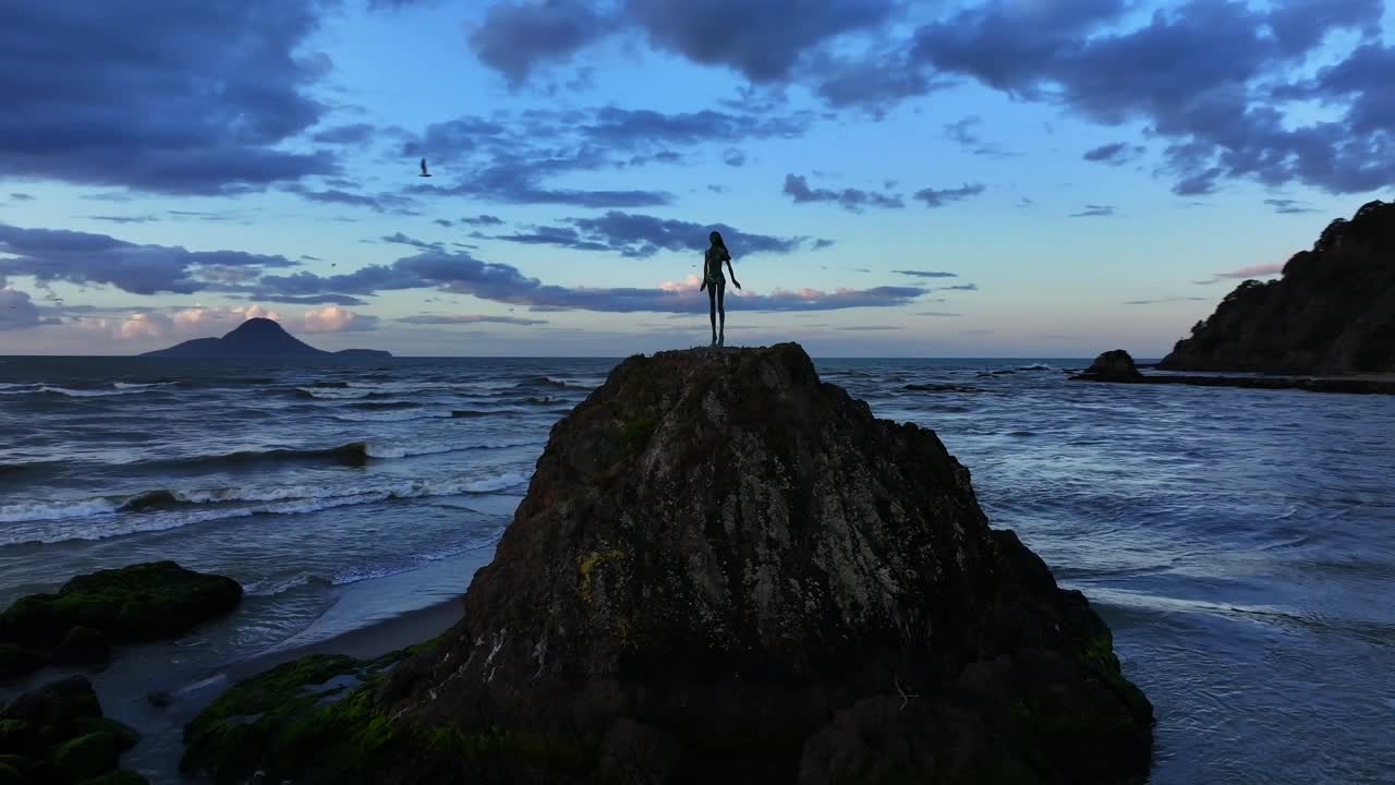 Sunset waves crash against a rocky outcrop, spray rising around a lone statue silhouetted in gold—still and solemn amid the ocean’s restless glow.