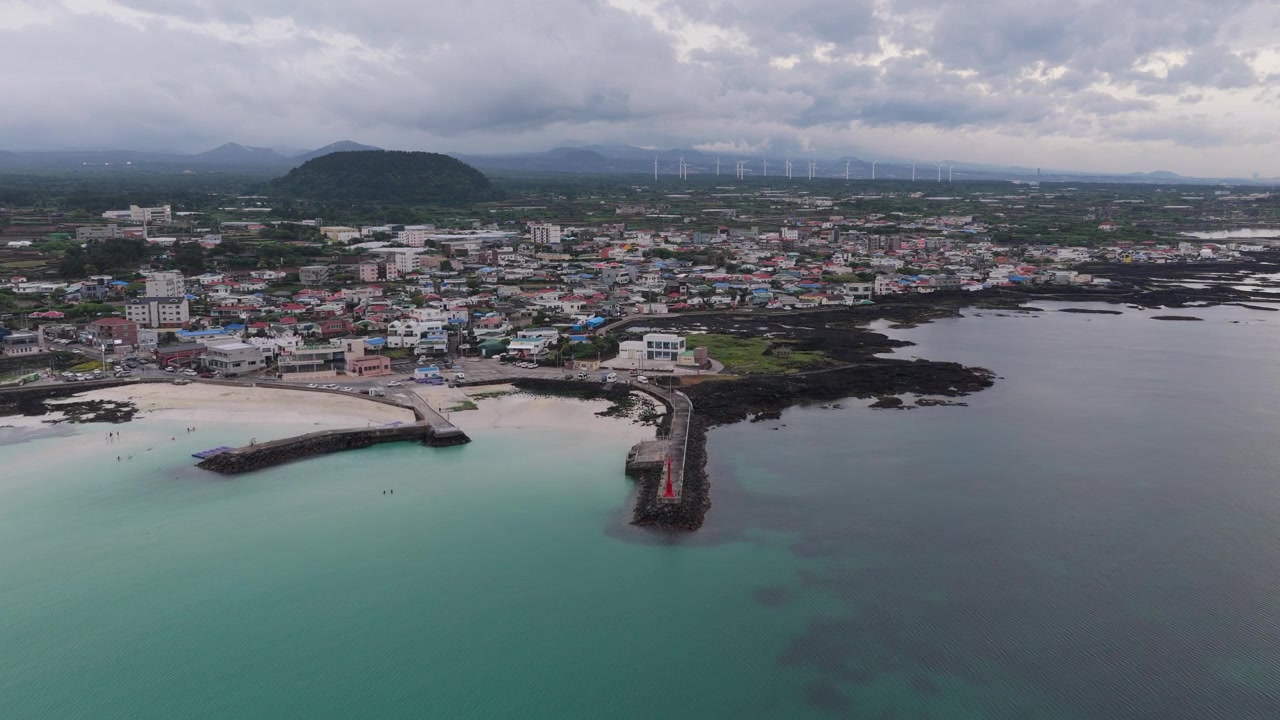 Cảnh quay ngang cận cảnh di chuyển tiến về phía trước trên Bãi biển Gimnyeong trên đảo Jeju, Hàn Quốc, hiển thị nước màu ngọc lam, bến cảng, bãi biển, làng mạc, đường bờ biển và đá núi lửa