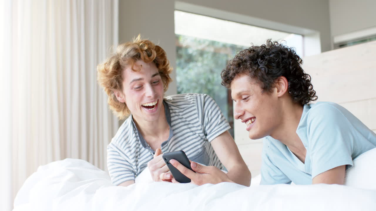 una pareja gay feliz y diversa acostada en la cama y usando un teléfono inteligente en casa, en cámara lenta