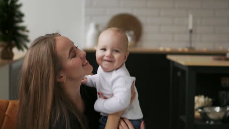 Mom-hugs-the-baby-and-the-child-smiles-looking-at-his-beloved-mother.-Together-stand-in-the-white-kitchen-on-Christmas-eve-on-the-background-of-garlands-and-Christmas-trees.-Happy-Mother-and-Baby-kissing-and-hugging.Maternity-concept-Motherhood-Beautiful-Happy-Family-Stock-Video-Footage-in-Slow-Moti