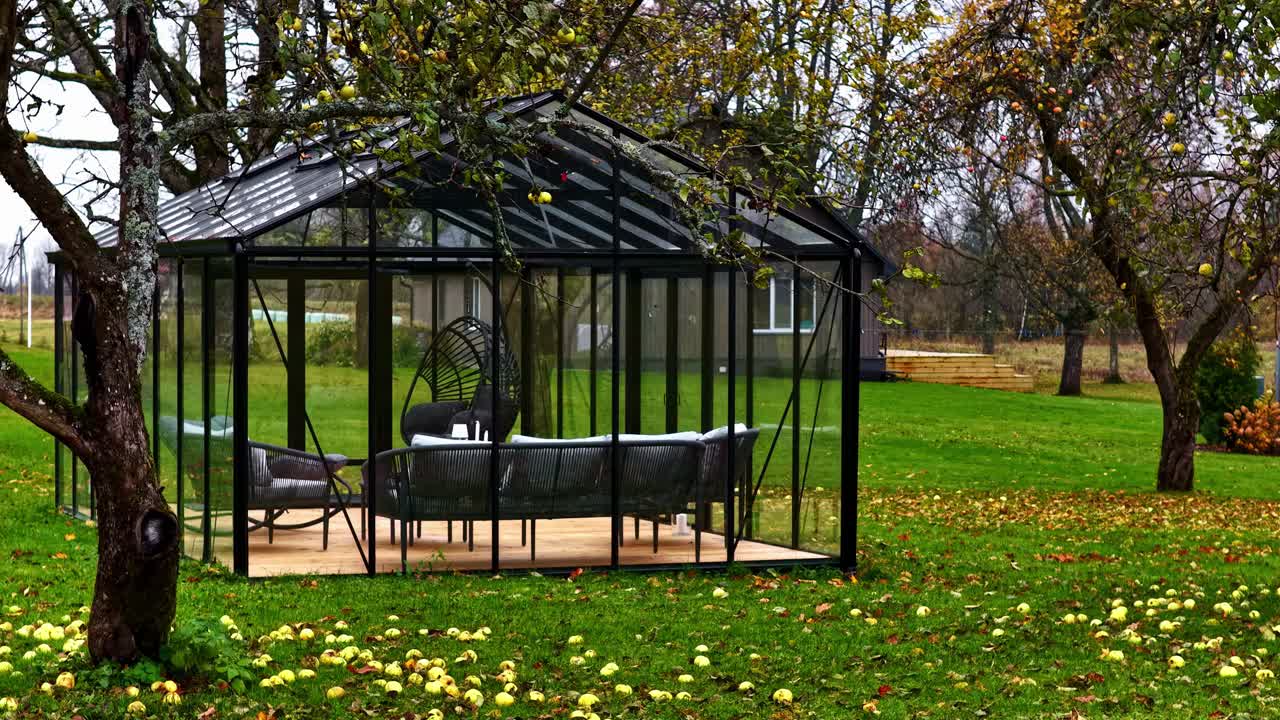 Modern black-framed glass conservatory furnished for relaxation among apple trees