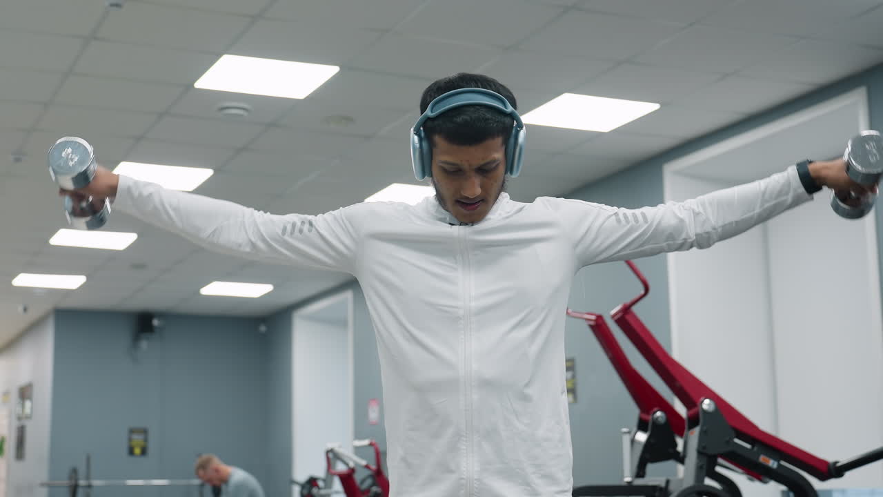 focused fitness coach raising dumbbells to shoulder height while training in gym, wearing wireless headphones and white zip-up top, with slightly blurred person seated in background