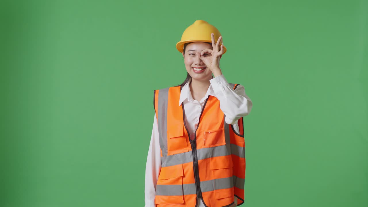 Asian Female Engineer With Safety Helmet Showing Ok Hand Sign Over Eye And Smiling To Camera While Standing In The Green Screen Background Studio