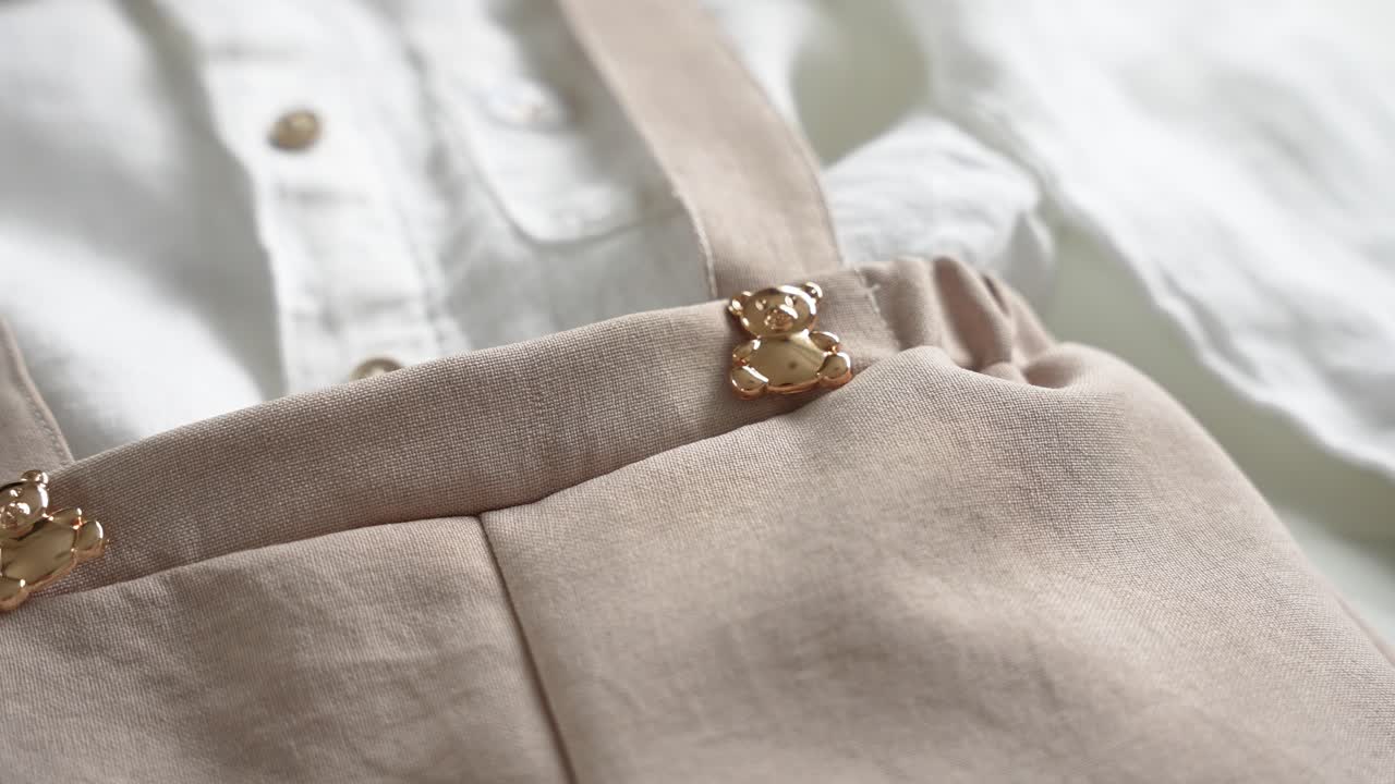 Close-up of beige baby suspenders with a cute golden teddy bear clasp detail, paired with a white linen shirt, styled for a christening, baptism, or special event