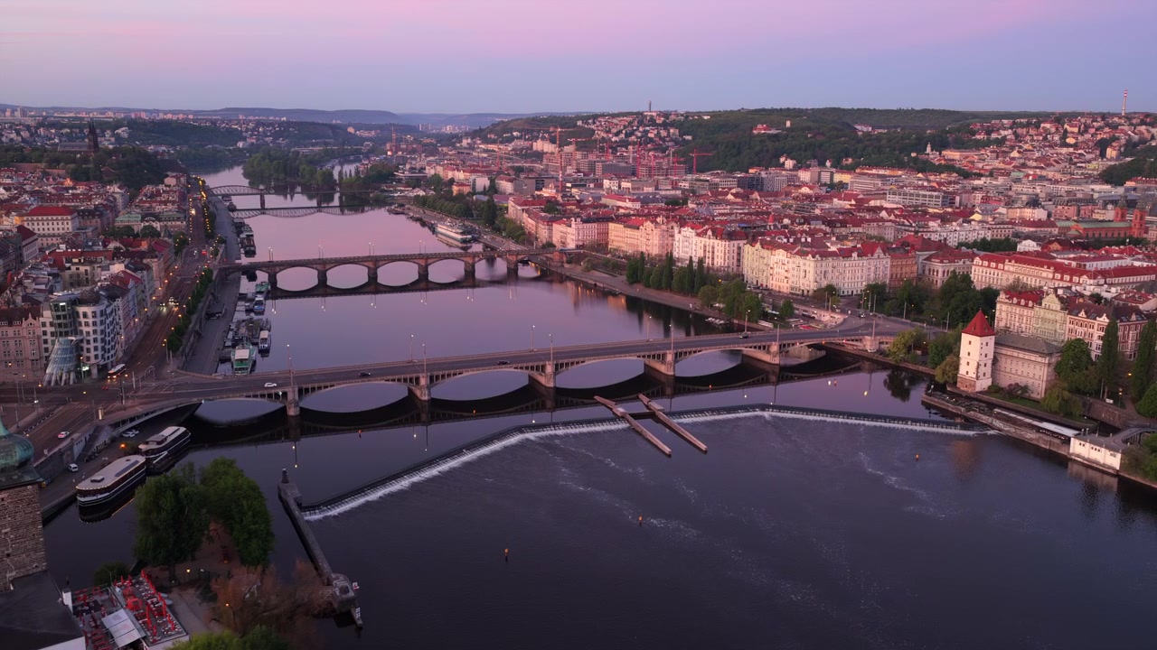 Vista aerea con drone del fiume Moldava all'alba viola-rosa, con diversi ponti, la Casa Danzante visibile in basso a sinistra, uno sbarramento d'acqua in primo piano e la città dai tetti rossi che si estende a destra, Praga.