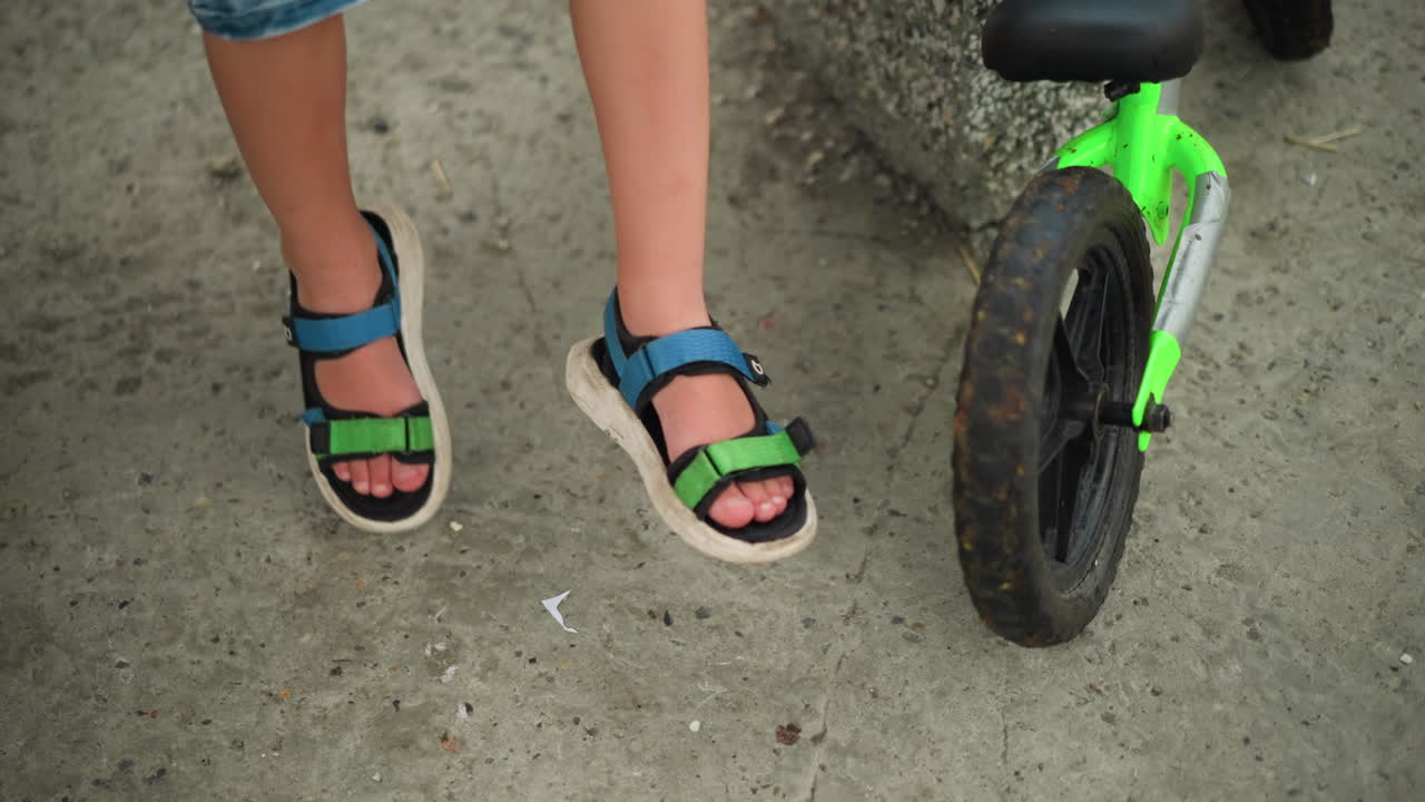 primer plano de un niño con sandalias azules y verdes, balanceando las piernas mientras está sentado, una bicicleta verde está estacionada cerca, y hay una vista parcial de la pierna de otro niño en el lado izquierdo