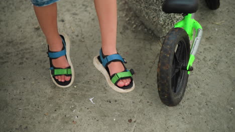 close-up of a child wearing blue and green sandals, swinging their legs while seated, a green bicycle is parked nearby, and there is a partial view of another child s leg on the left side