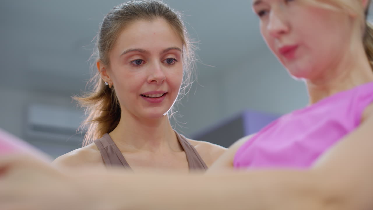 갈색 옷을 입은 강사는 분홍색 옷를 입은 여성이 앉아서 앉아있는 것을 면밀히 관찰하고 안내합니다. 여성의 팔은 움직이고 밝은 빛이 뒤에서 빛나며 피트니스 스튜디오에서 강사의 머리를 둘러싸고 반사됩니다.