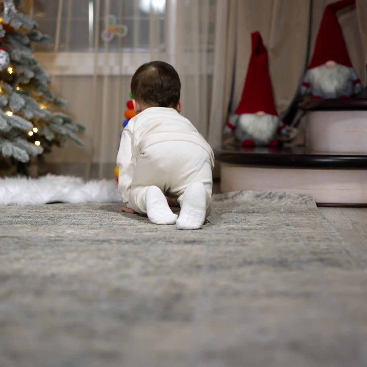 Little child in white clothes crawls by the room followed by camera. Baby stops at the toy pyramid near the Christmas tree
