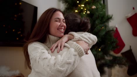 Happy-couple-guy-and-girl-sitting-opposite-each-other-and-hugging-in-white-sweaters-near-the-Christmas-tree-in-a-cozy-room-in-winter.-The-magic-of-Christmas-warmth-and-comfort-as-well-as-the-family-atmosphere-in-winter