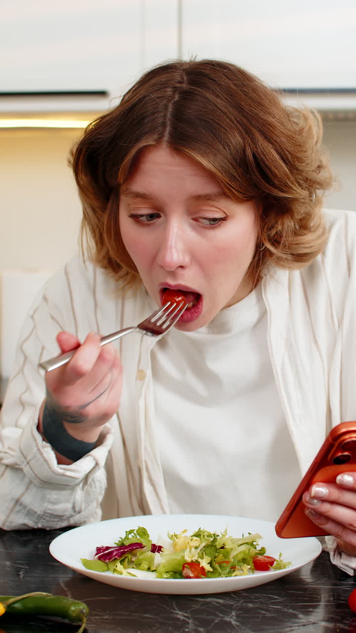 Mujer joven revisando el smartphone mientras come ensalada fresca en la cocina durante su pausa para el almuerzo de dieta saludable.