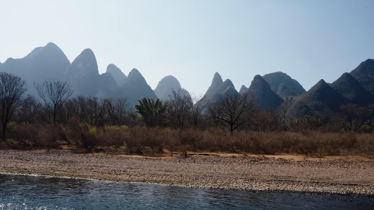 A row of sharp karst peaks rises above dry brush and rocky riverbanks along the Li River
